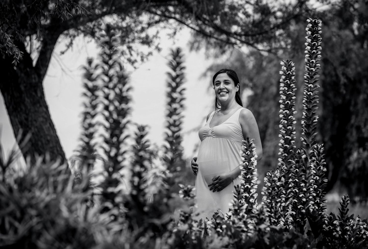 Black and white photo of a smiling pregnant woman in a garden surrounded by tall plants and trees.