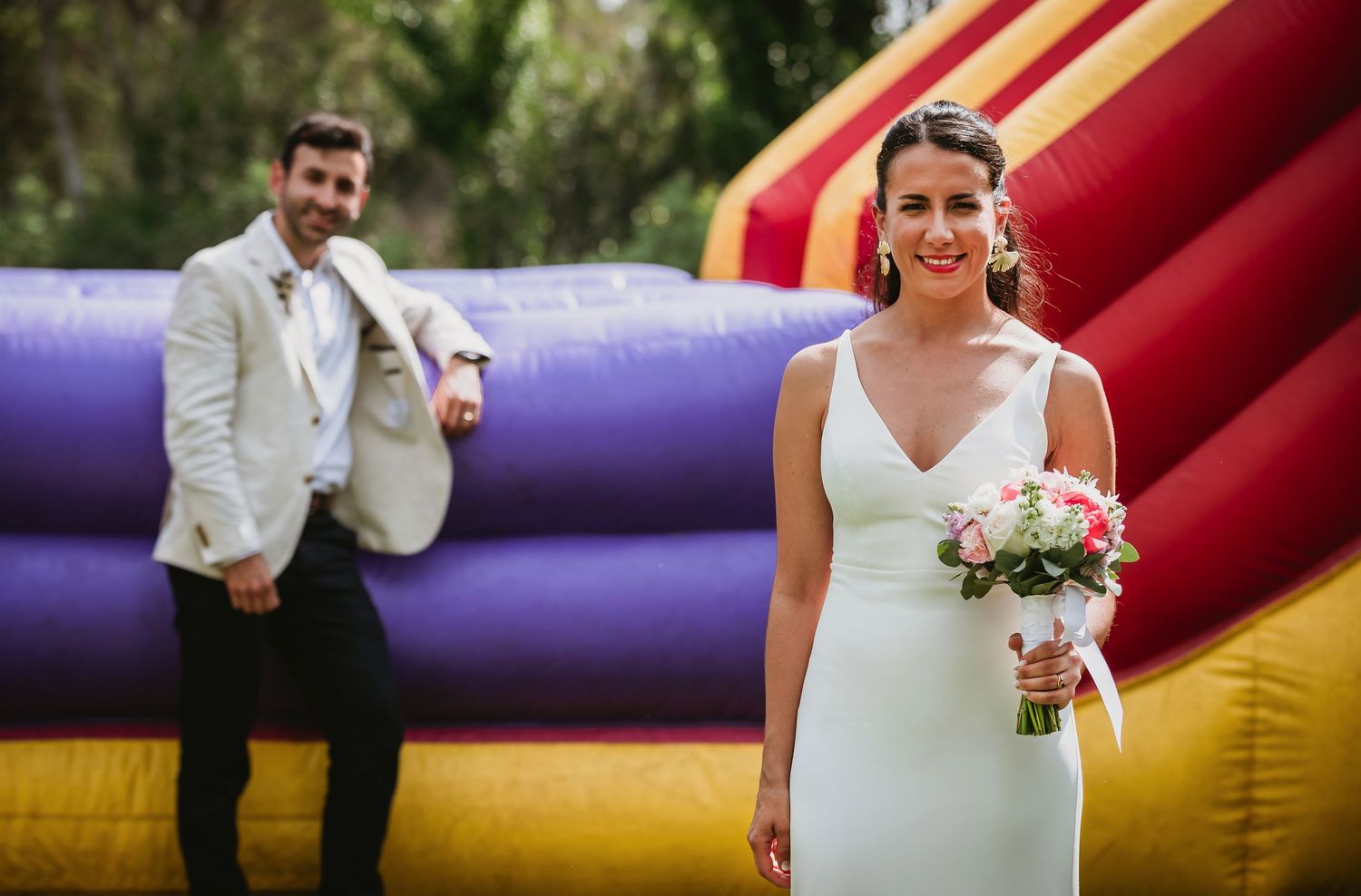 Bride in white gown holds pink bouquet while groom poses playfully near colorful bouncy castle at outdoor wedding.