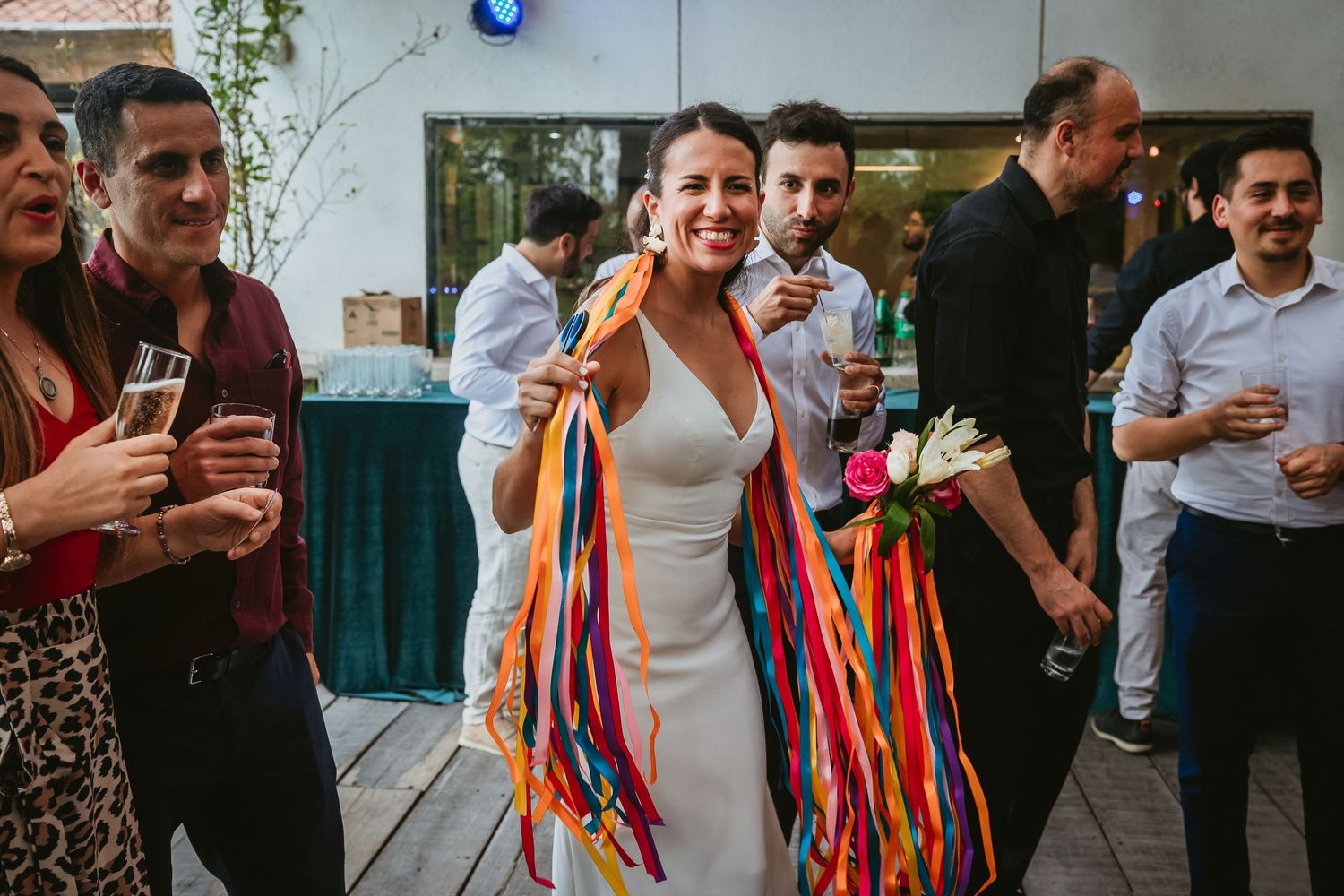 Bride in white dress with colorful ribbons smiles at wedding reception surrounded by guests.