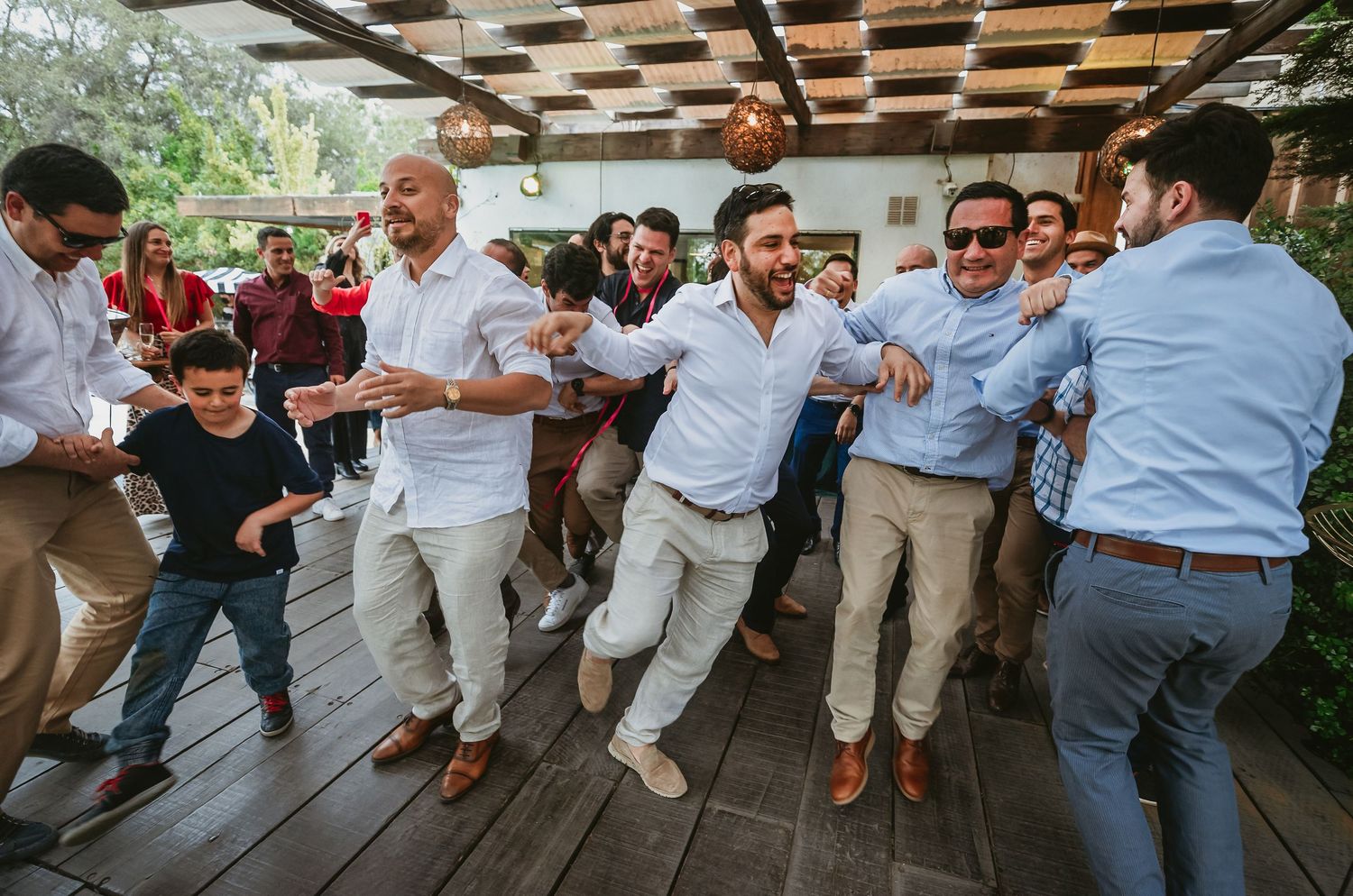 Men dancing energetically at an outdoor wedding reception on a wooden deck, with guests celebrating in the background.