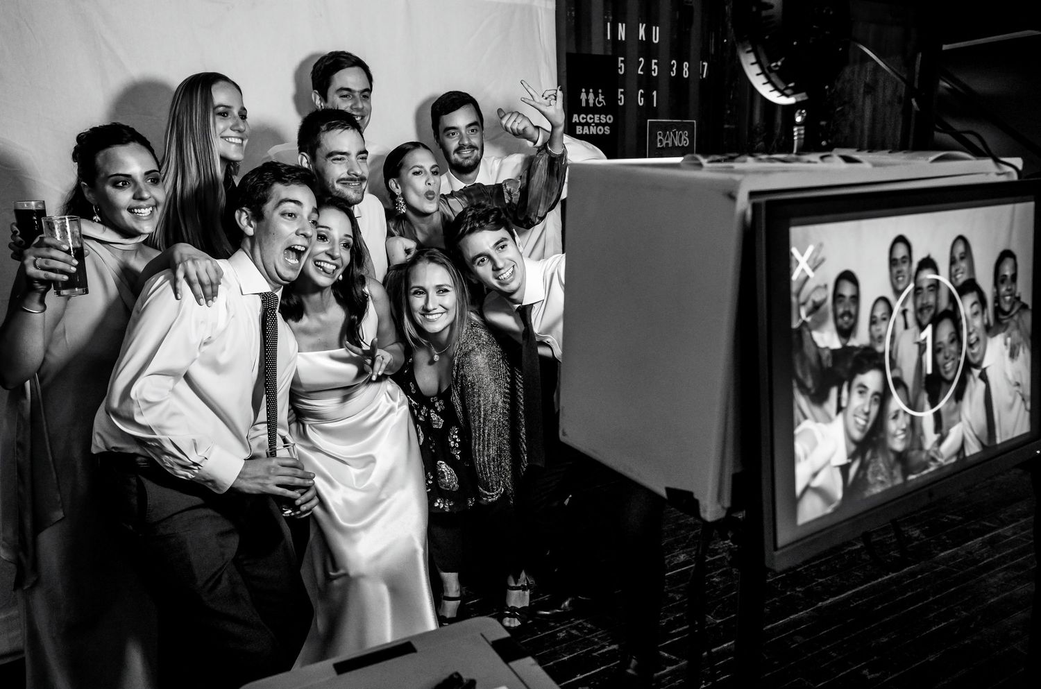 Wedding group selfie at photo booth, bride and groom with guests posing in black and white photo.
