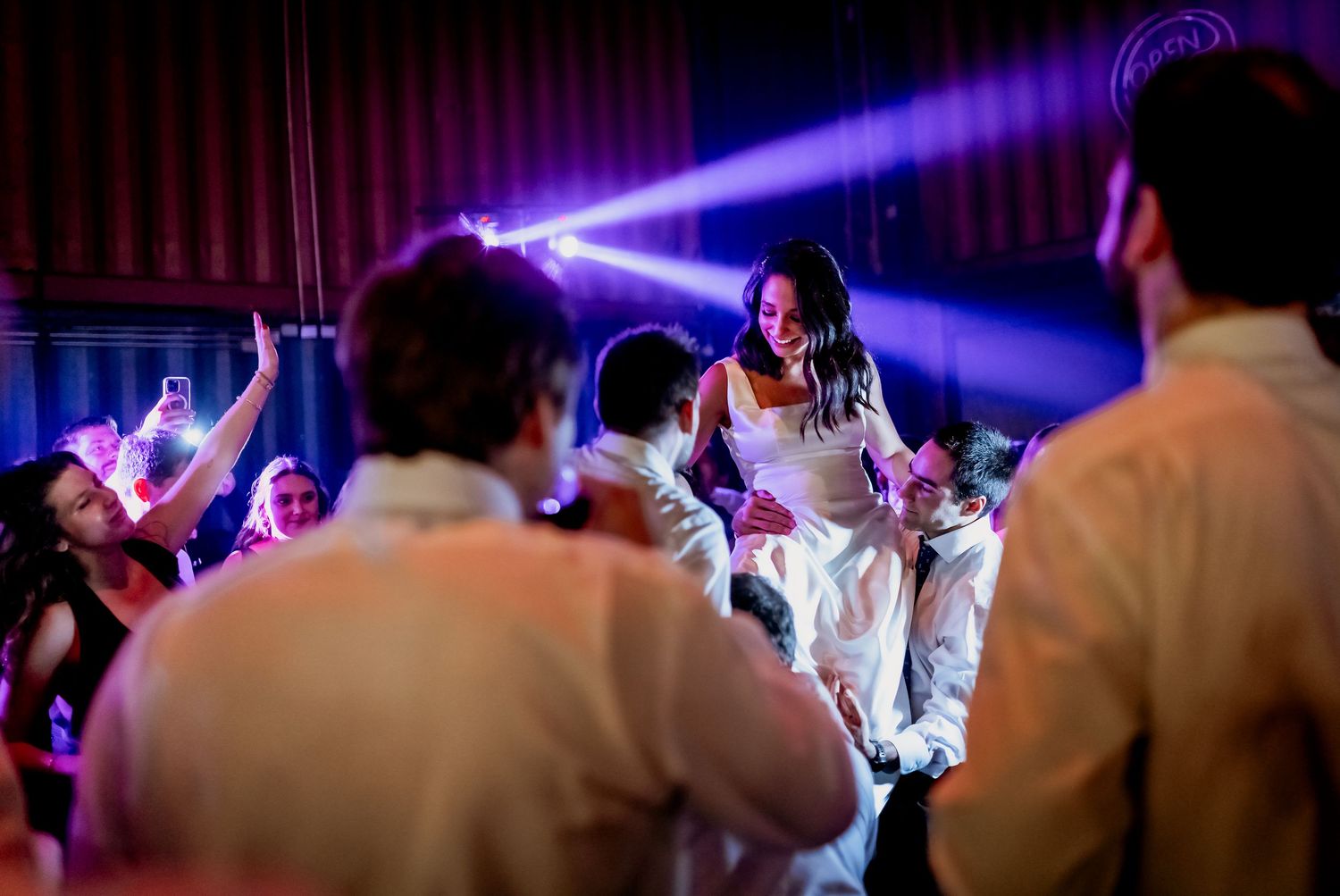 Bride being lifted by guests on dance floor at vibrant wedding reception party.