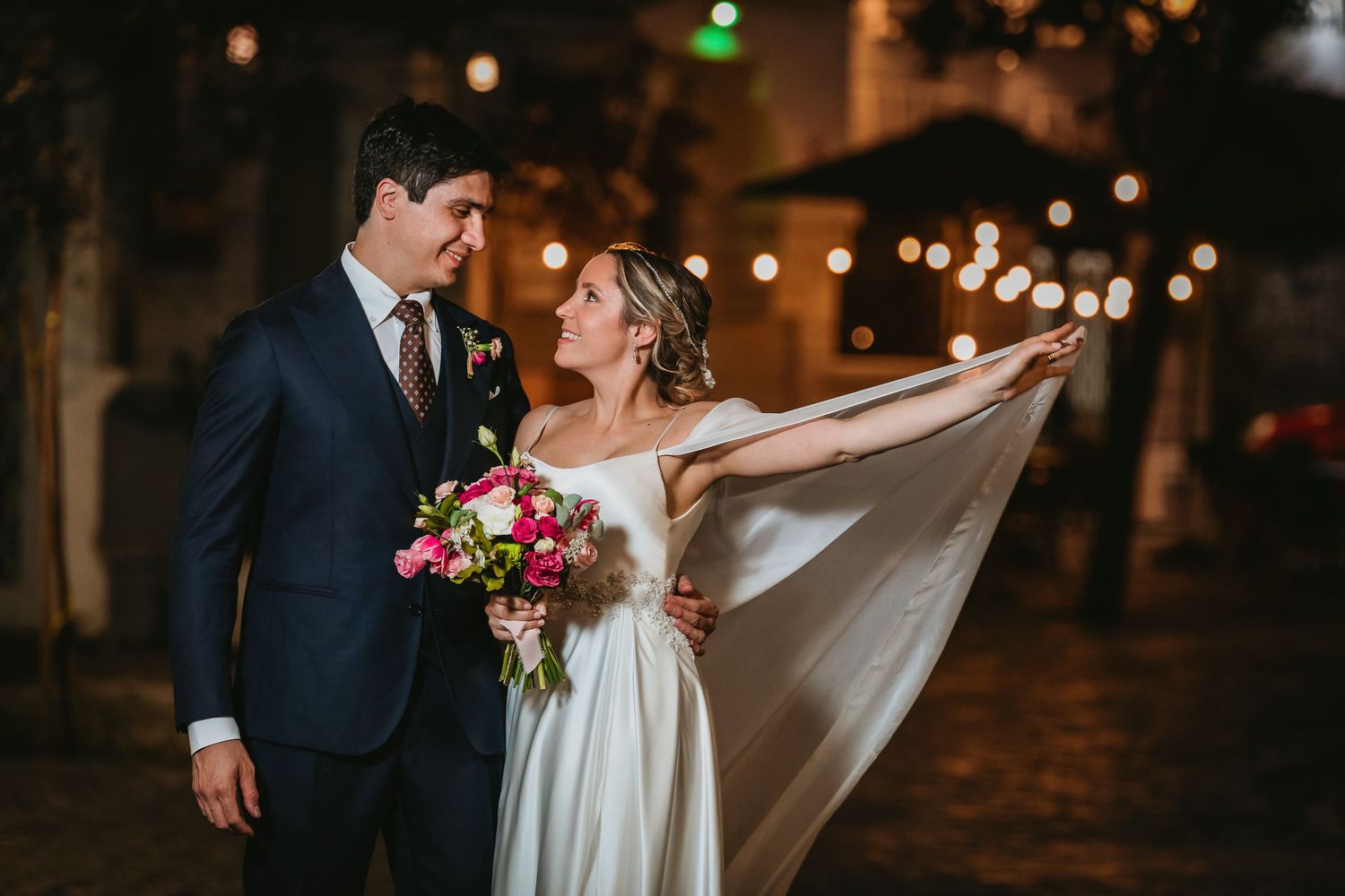Bride extending flowing white veil while groom watches lovingly under string lights at night.