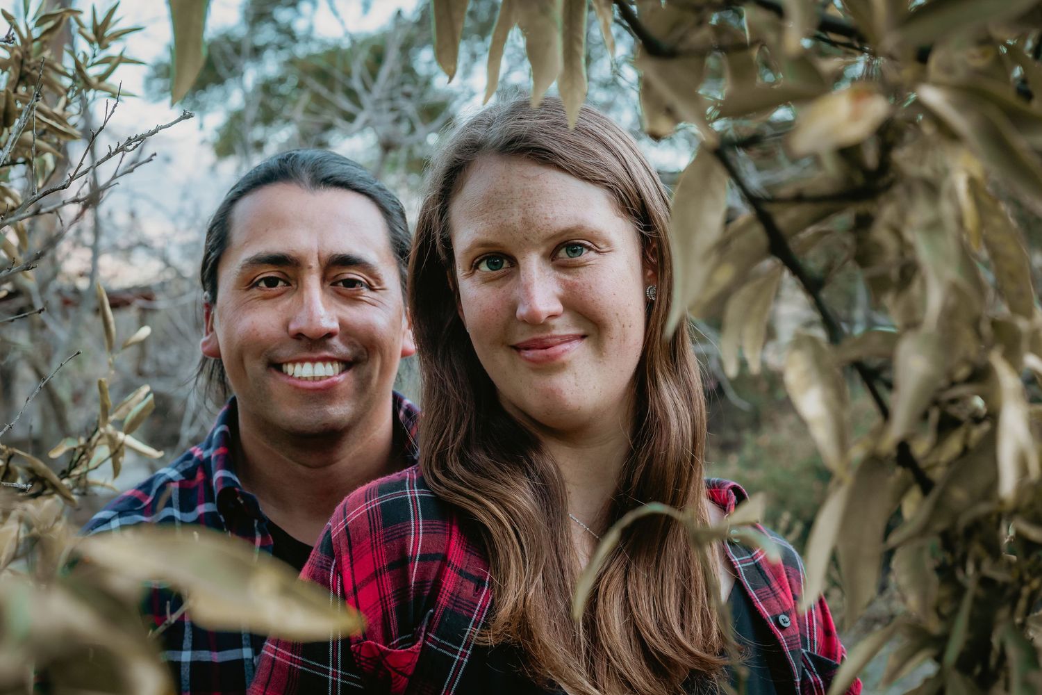 A close-up of a smiling couple peeking through autumn foliage branches in a natural outdoor setting.