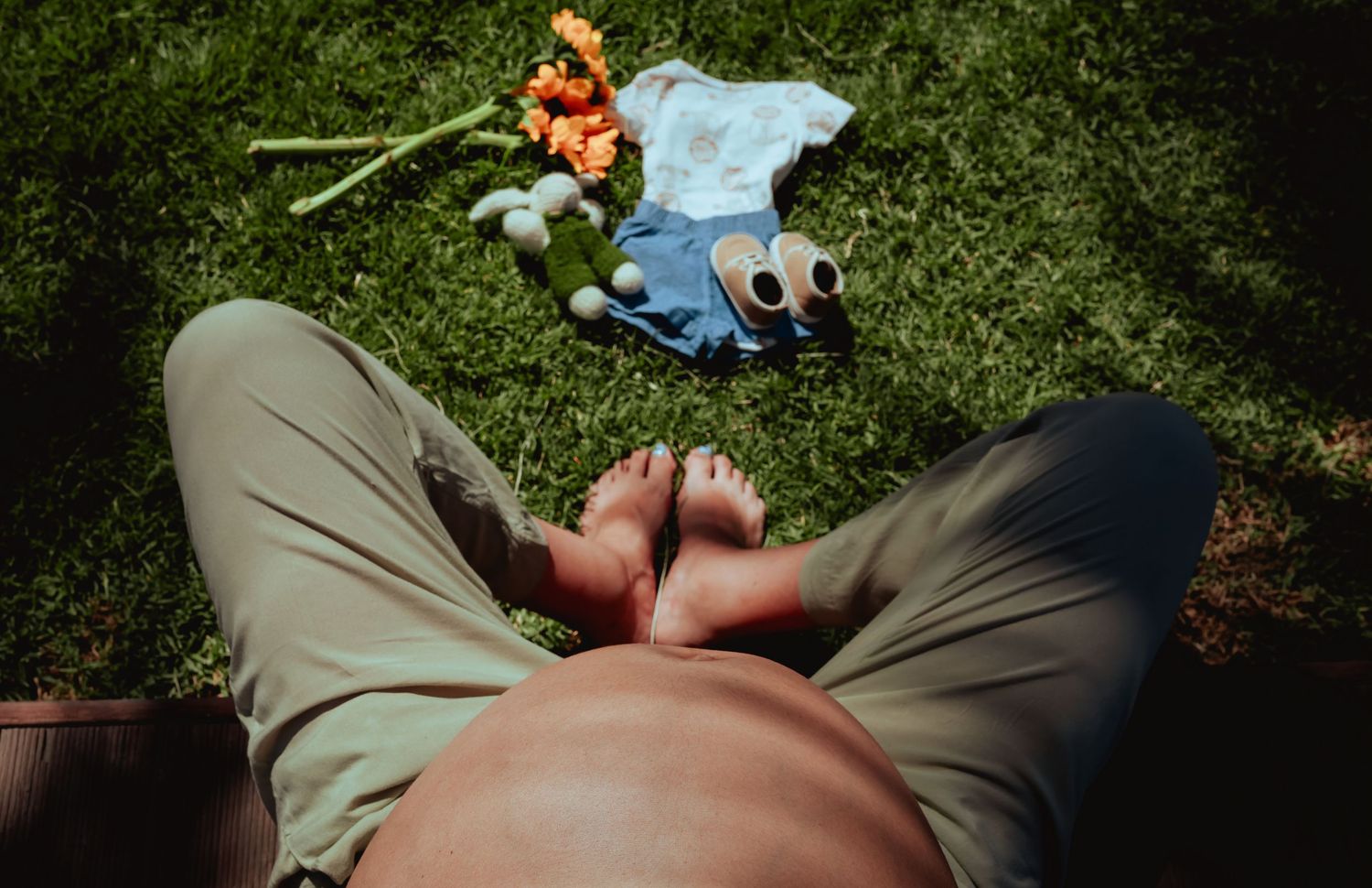 Overhead view of pregnant woman resting on grass with baby clothes, toys, and flowers beside her.