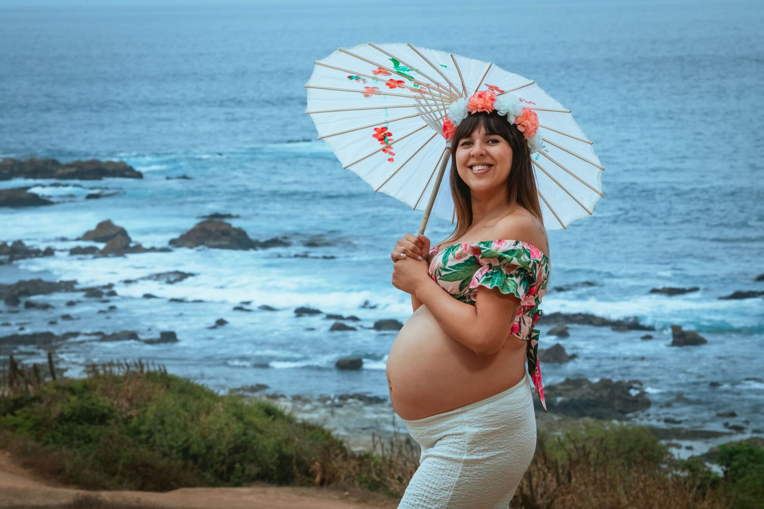 Pregnant woman holding parasol umbrella with floral crown by ocean.
