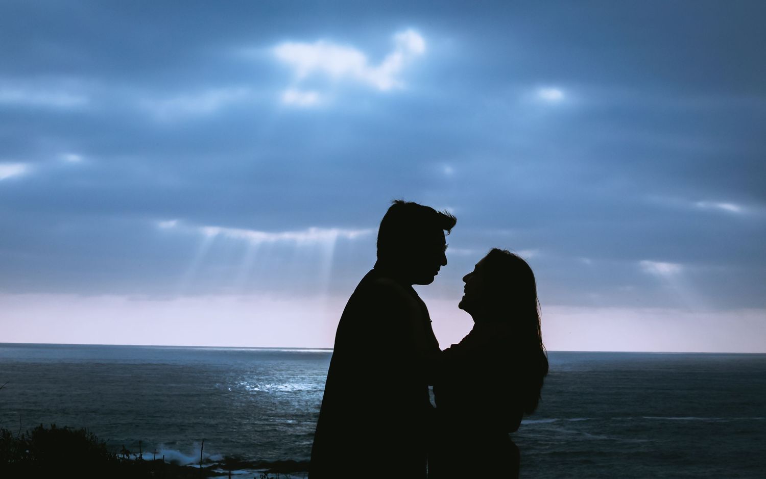 Silhouette of a romantic couple facing each other by the ocean under dramatic cloudy skies with sunbeams breaking through.
