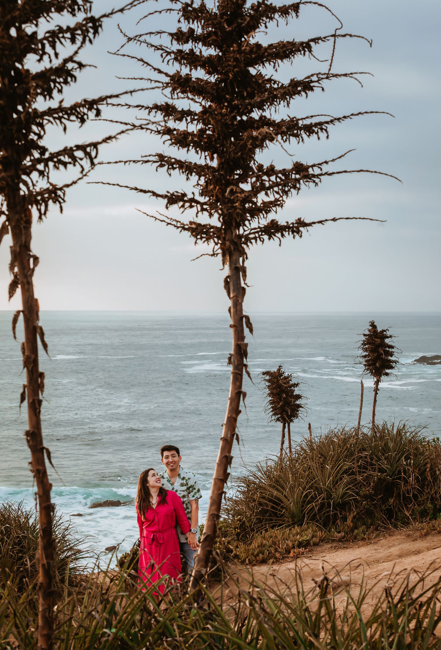 Couple standing together on a coastal cliff path surrounded by tall trees with ocean waves in the background.