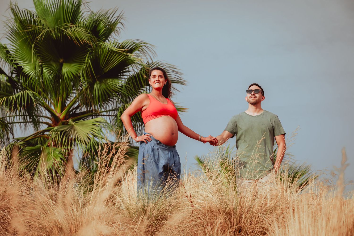 Pregnant woman in red crop top and man holding hands walking through golden grass near palm tree.