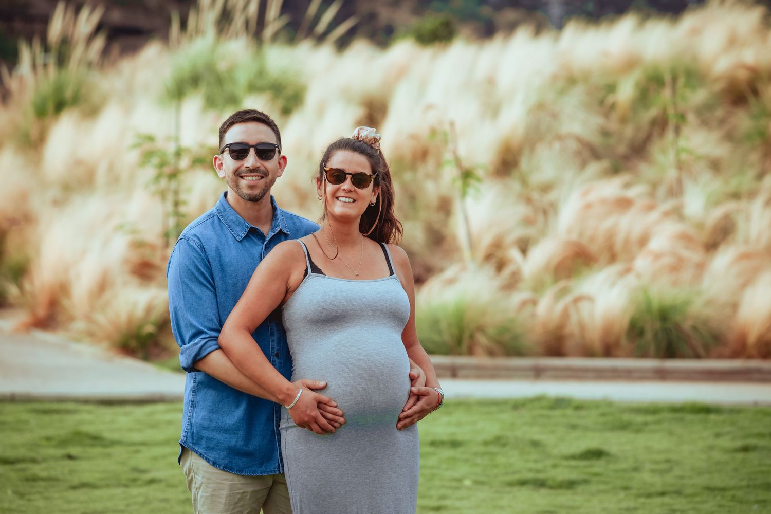 Smiling couple posing outdoors, pregnant woman in grey dress cradling baby bump with partner in blue shirt.