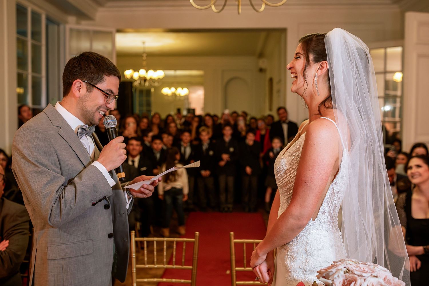 Groom reads vows to smiling bride in wedding gown and veil during elegant indoor ceremony with guests.