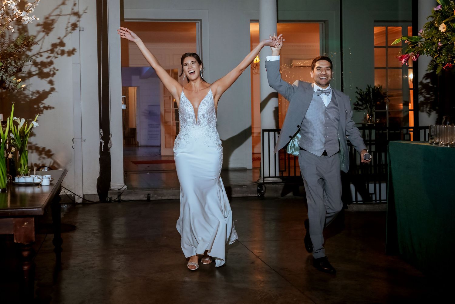 Bride in white gown and groom in gray suit celebrate joyfully at their wedding reception entrance.