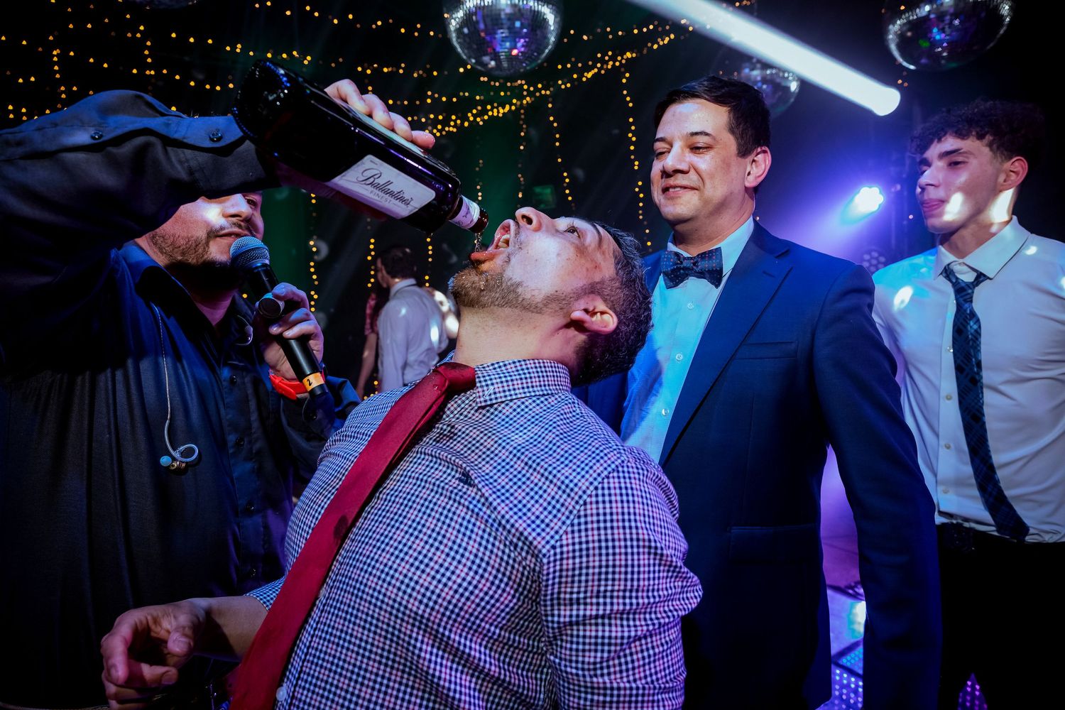 Men in formal attire at a party, one drinking from a bottle while others watch under disco lights.