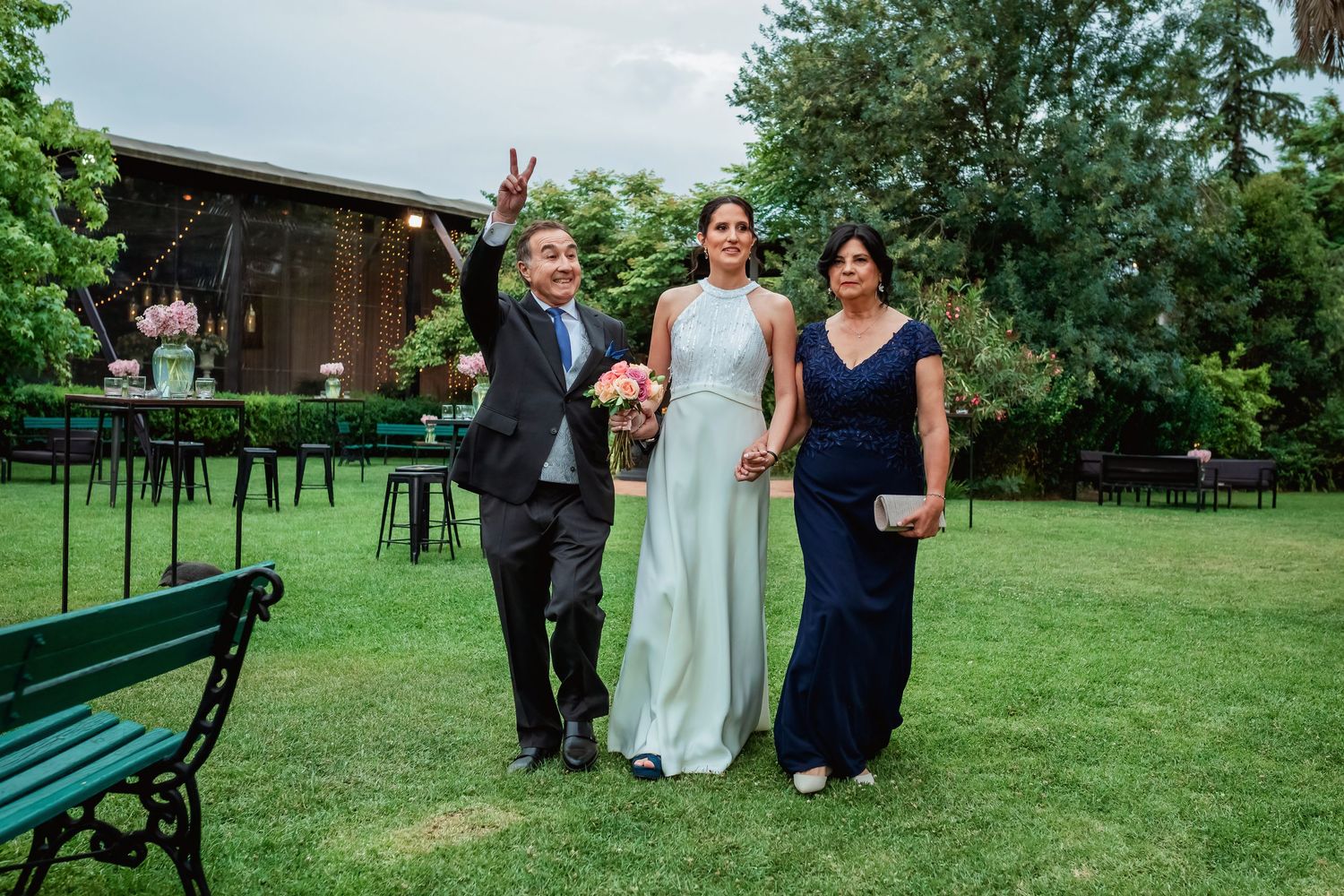Bride in white gown walks with parents across green lawn at outdoor wedding venue with garden benches.
