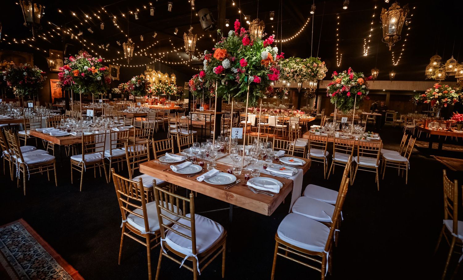 Elegant wedding reception hall with tall floral centerpieces, string lights, and gold chiavari chairs.