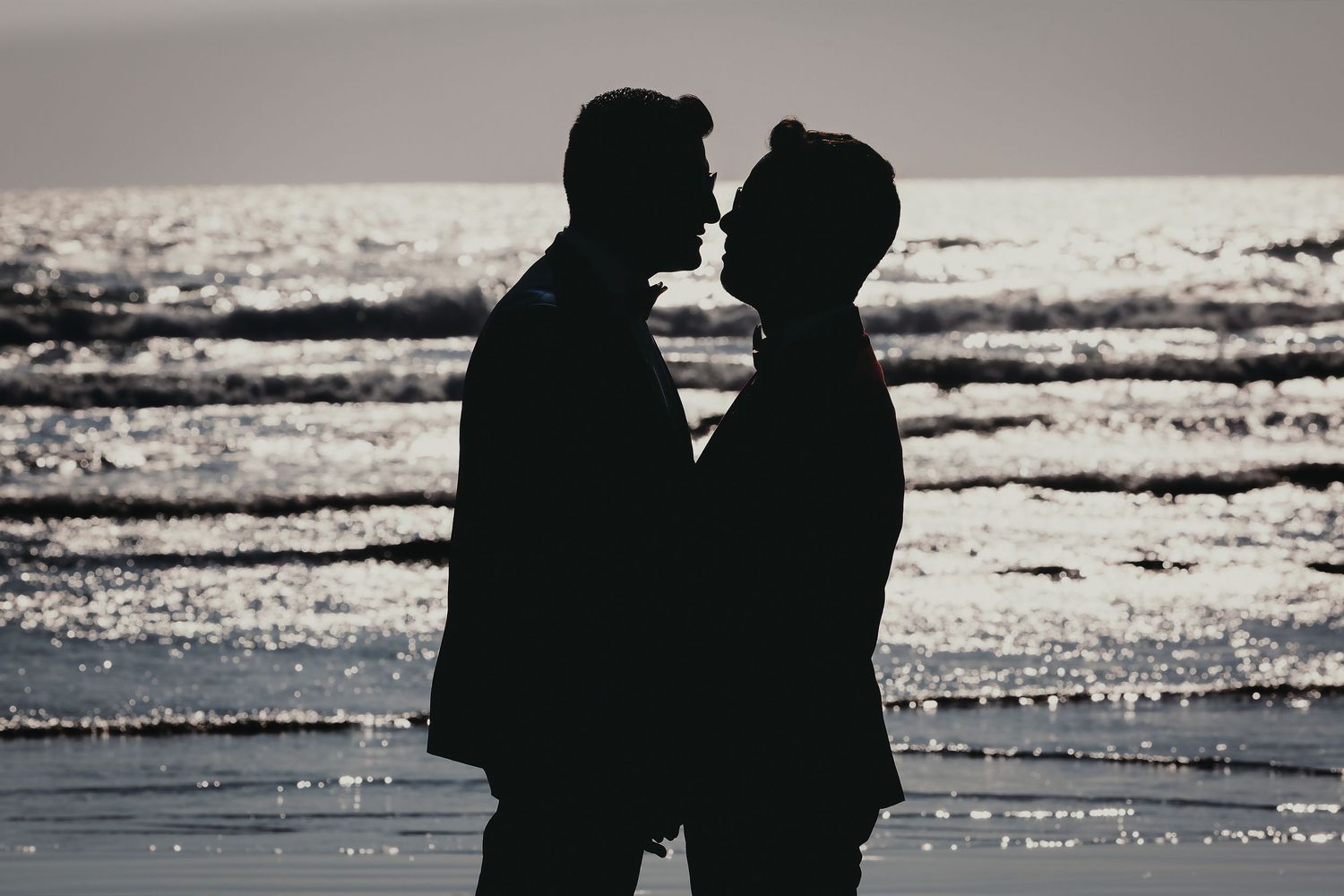 Silhouette of two grooms about to kiss on a sparkling beach at golden hour.