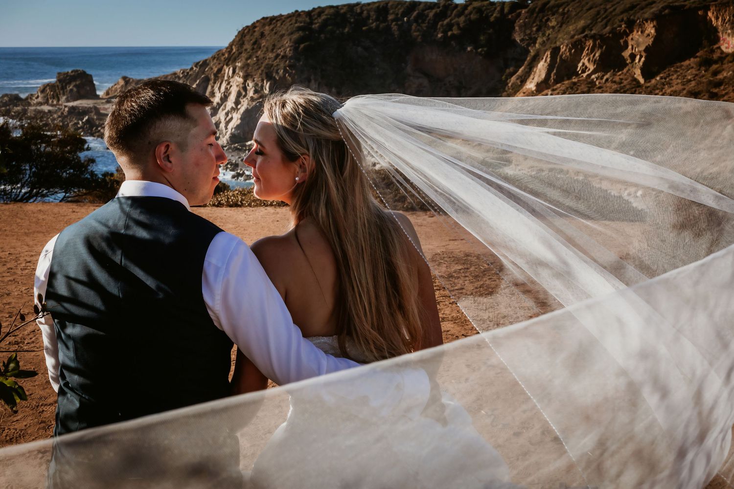 Bride and groom share an intimate glance while seated outdoors as her sheer veil billows around them by the coast.