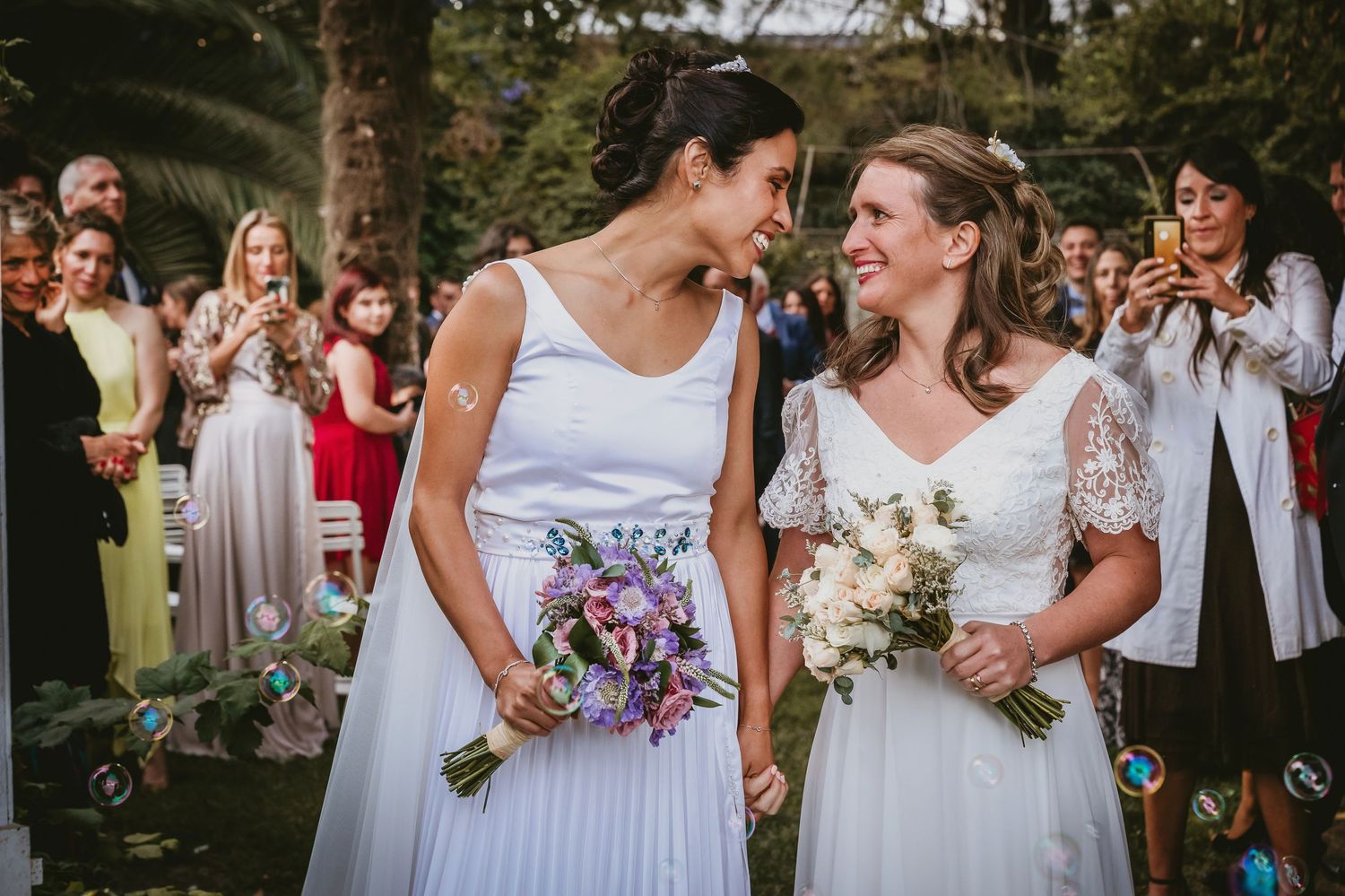 Two brides in white dresses share a joyful moment walking down the aisle surrounded by guests blowing bubbles.
