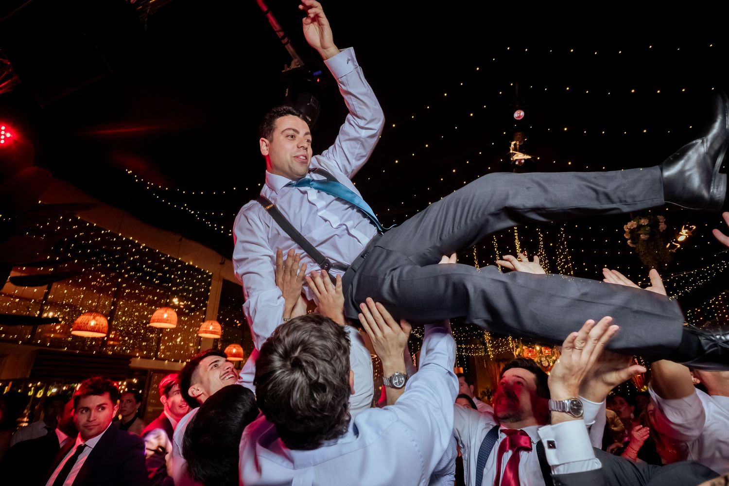 Groom is crowd-surfed by cheering guests at a lively wedding reception under string lights.
