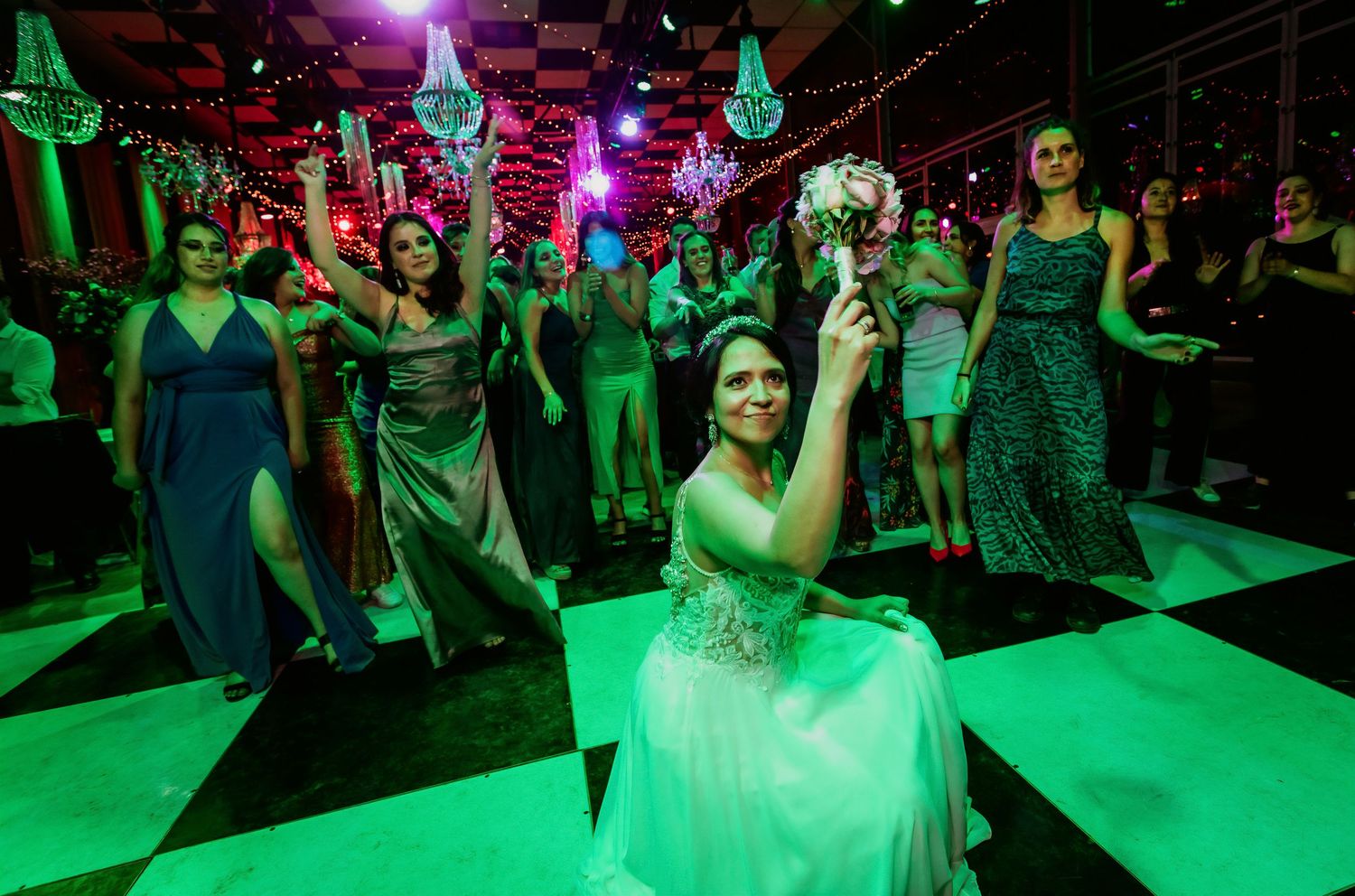 Bride kneels holding bouquet preparing for bouquet toss at wedding reception on checkered dance floor.
