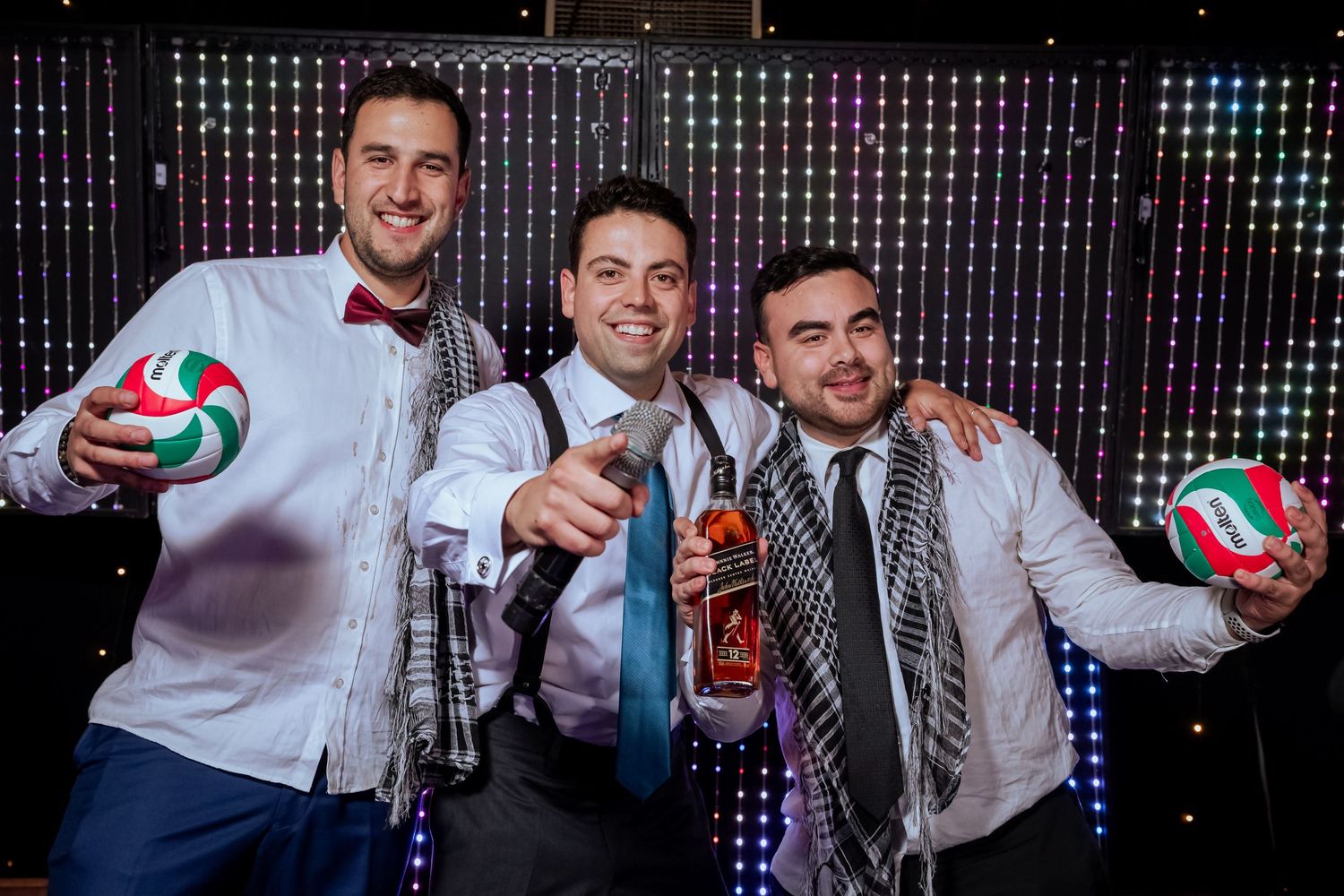 Three men in white shirts pose with Molten volleyballs and whiskey bottle at wedding.