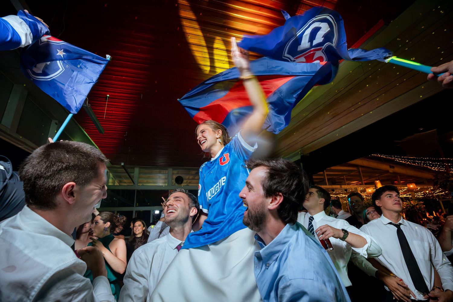 Wedding guest lifted on shoulders waves blue flag among celebrating crowd at reception.