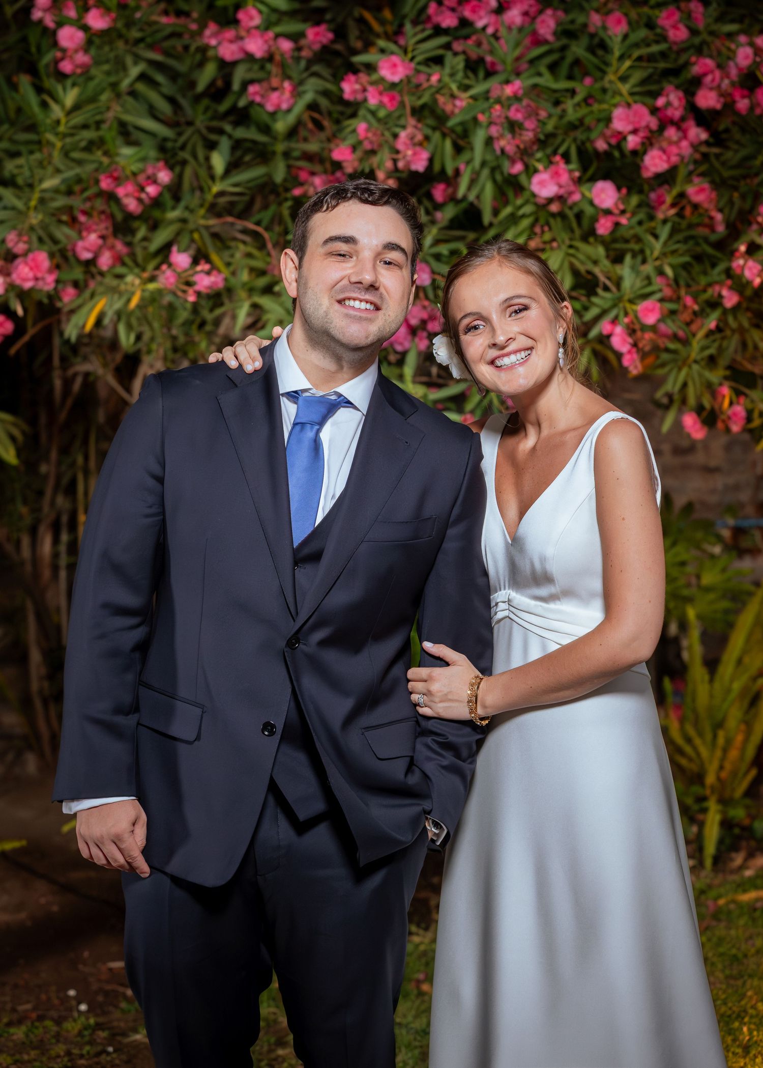 Smiling newlywed couple poses together in front of blooming pink oleander flowers at night.