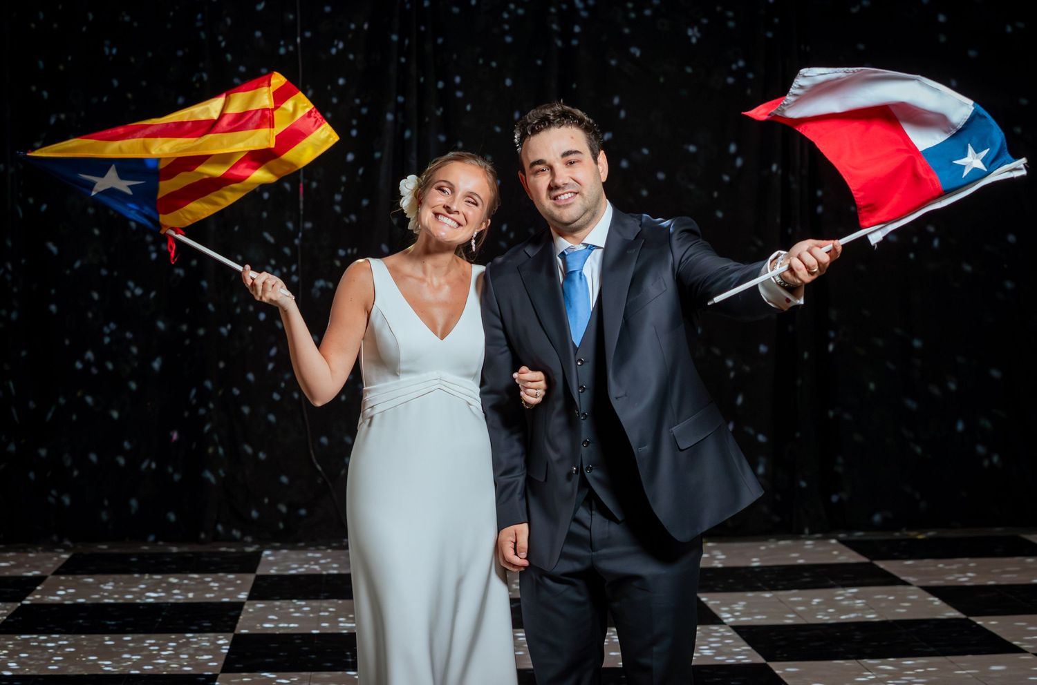 Bride and groom smiling and waving Catalan and Chilean flags at their wedding reception on a checkered dance floor.