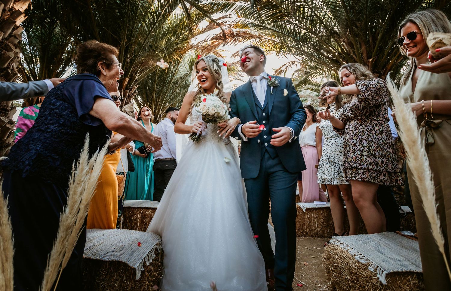 Bride and groom exit ceremony through pampas grass aisle lined with cheering guests outdoors.