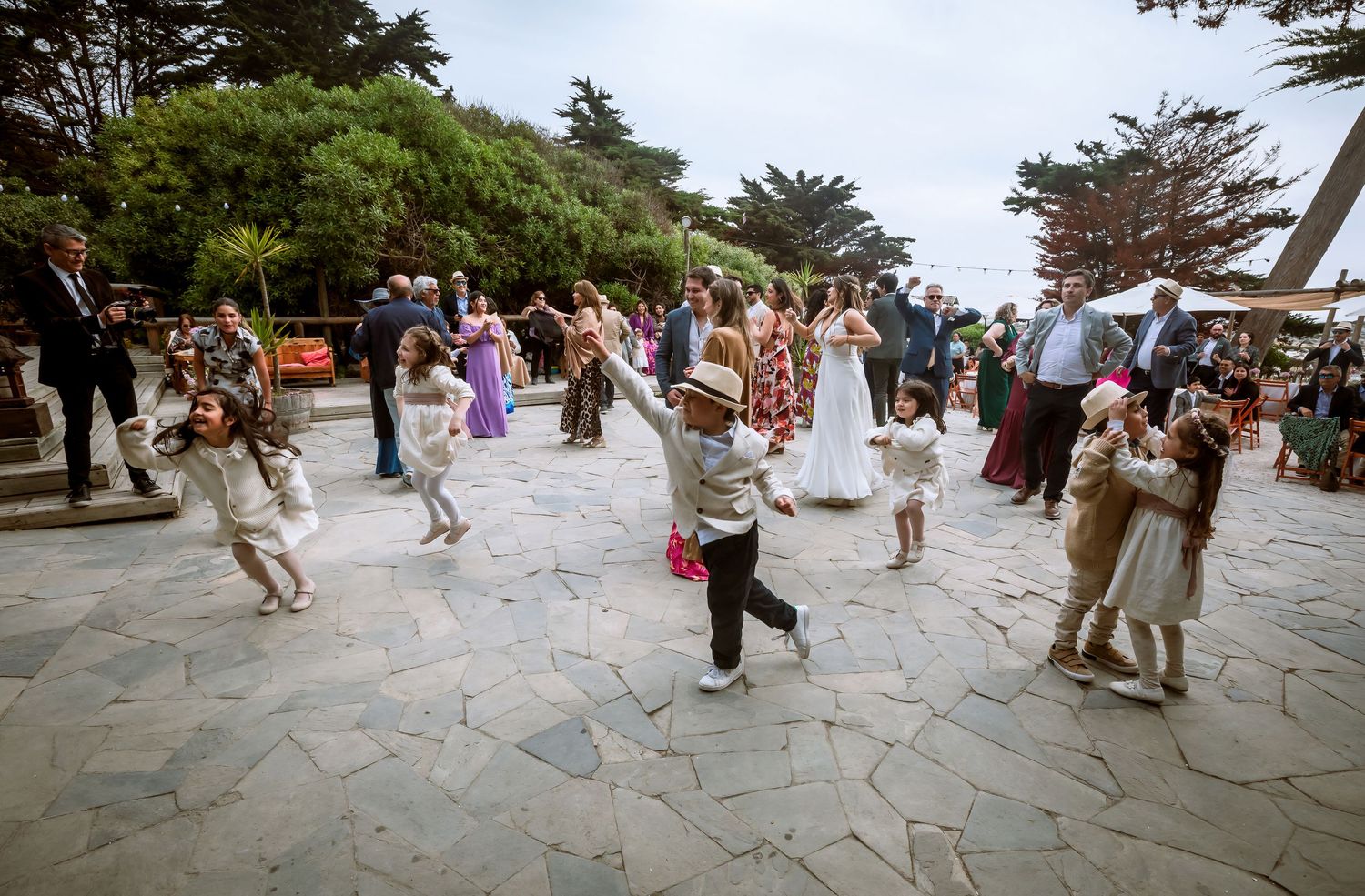 Children dancing joyfully at an outdoor wedding reception on a stone patio surrounded by guests.