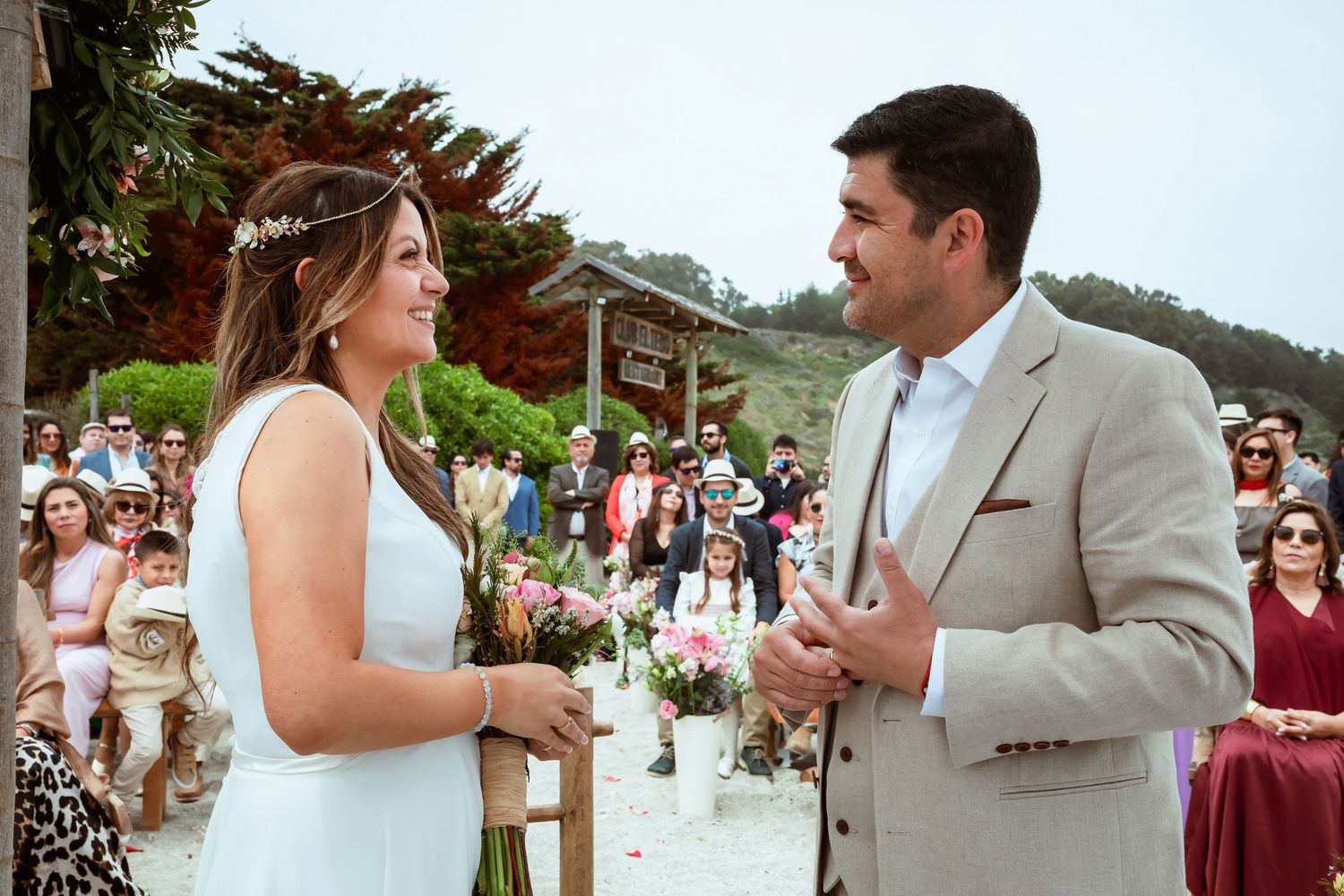 Bride and groom exchange vows at an outdoor beach wedding ceremony with guests seated behind them.