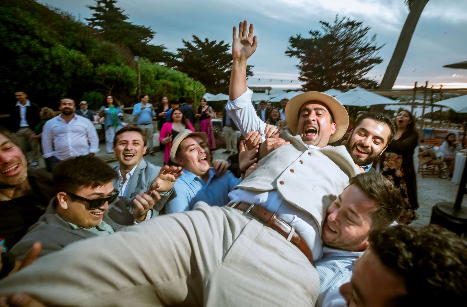 Groom crowd-surfed by cheering groomsmen and guests at lively outdoor wedding reception.