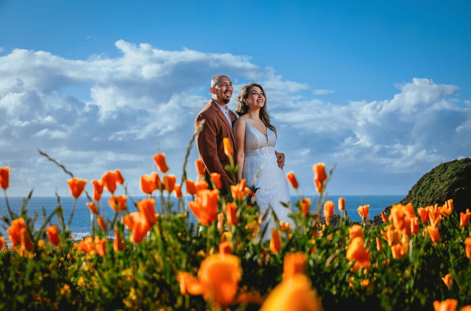 Couple in wedding attire embracing behind vibrant orange poppies on a coastal hillside under a bright blue sky with white clouds.