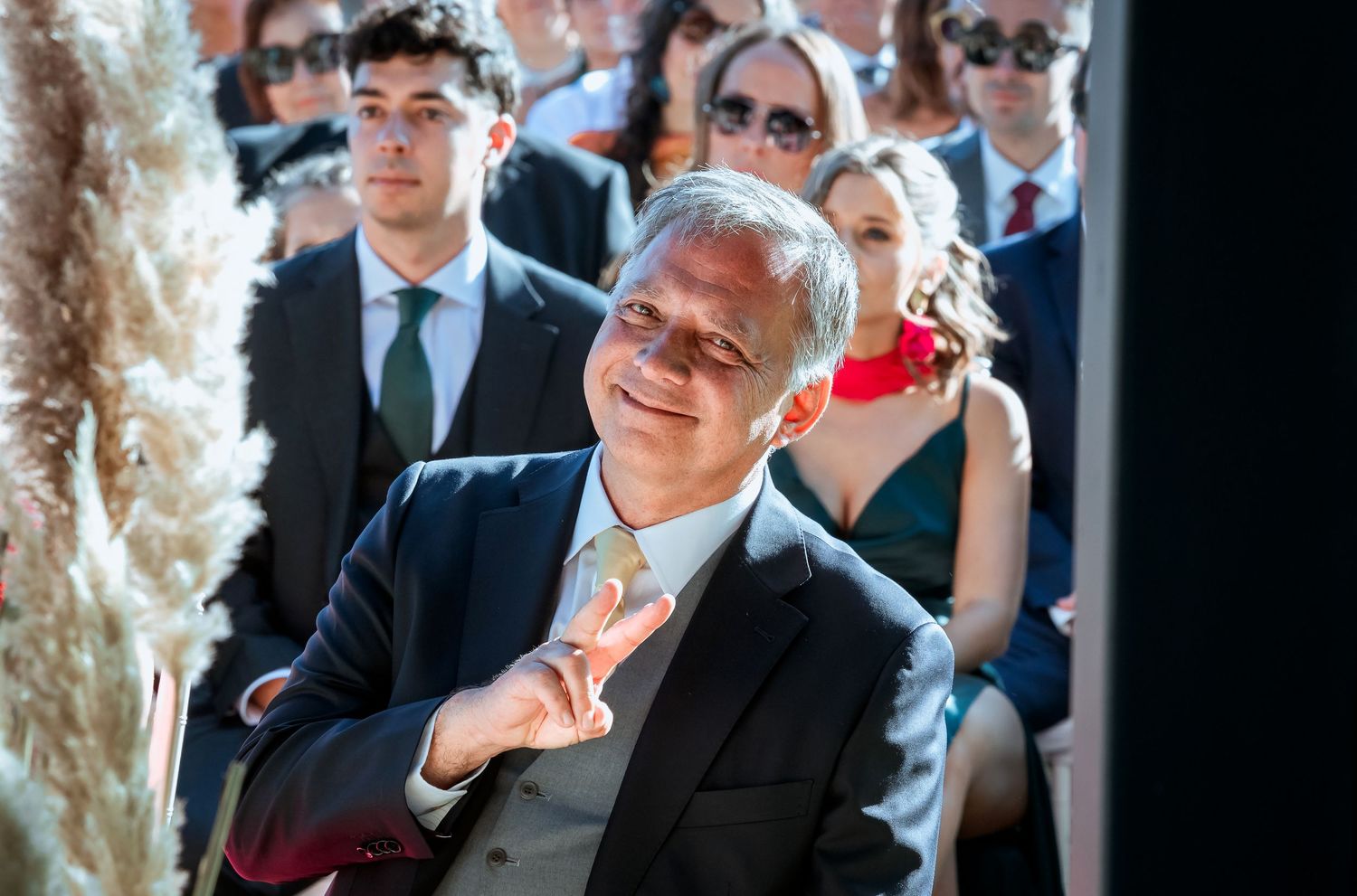 Elegant wedding guest in a suit gesturing and smiling during an outdoor wedding ceremony.