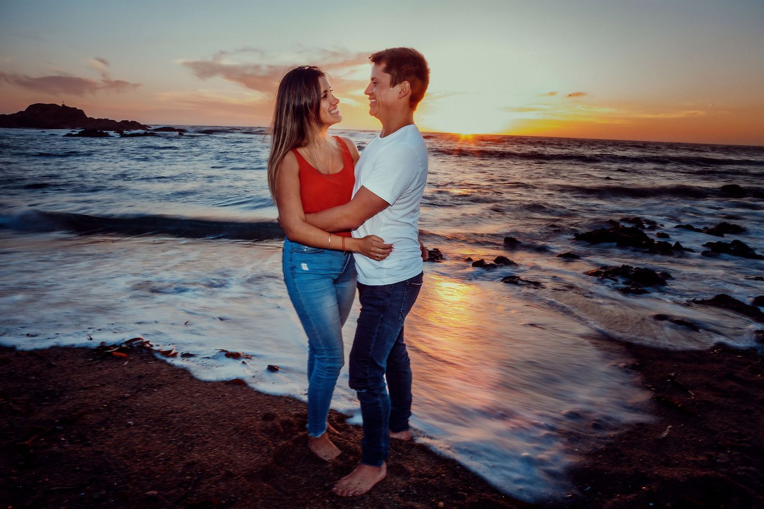 Couple holding each other close on a rocky beach shore during a vibrant romantic sunset.