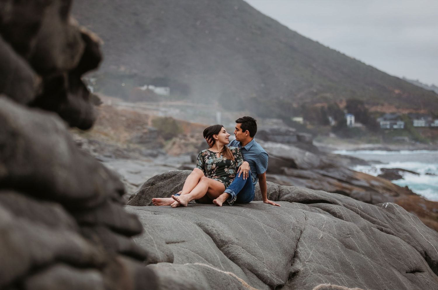 Couple sitting intimately on coastal rocks with ocean waves and mountain backdrop.