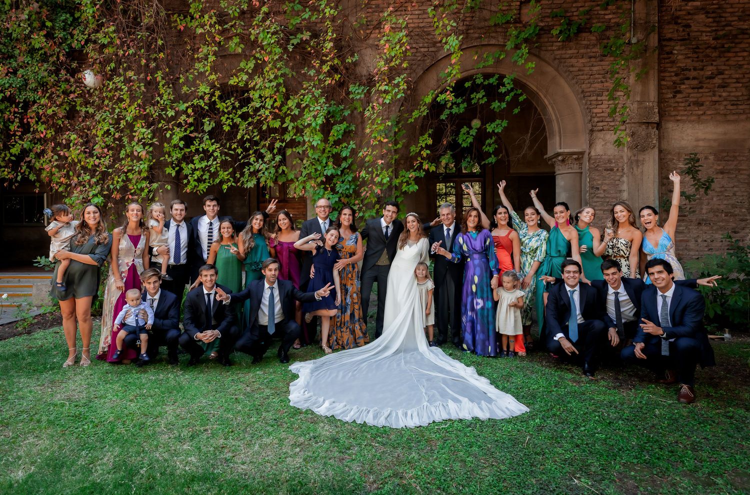 Large wedding party group photo with bride and groom posing on green lawn before ivy-covered arch.