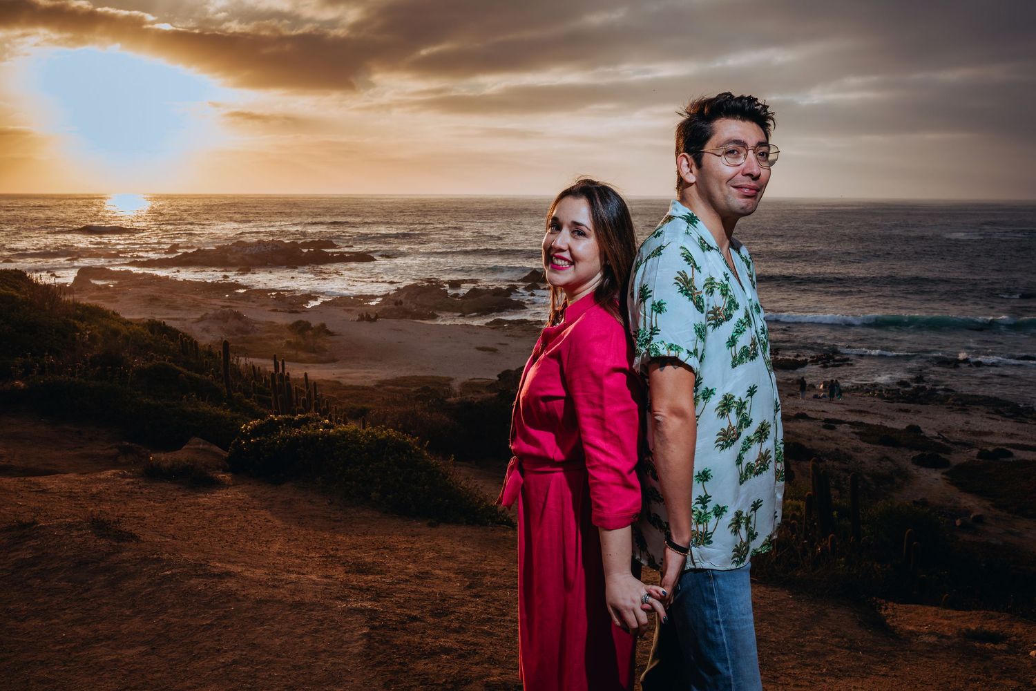 Couple holding hands at sunset on coastal cliffs, woman in red dress smiling, man in floral shirt, ocean backdrop.