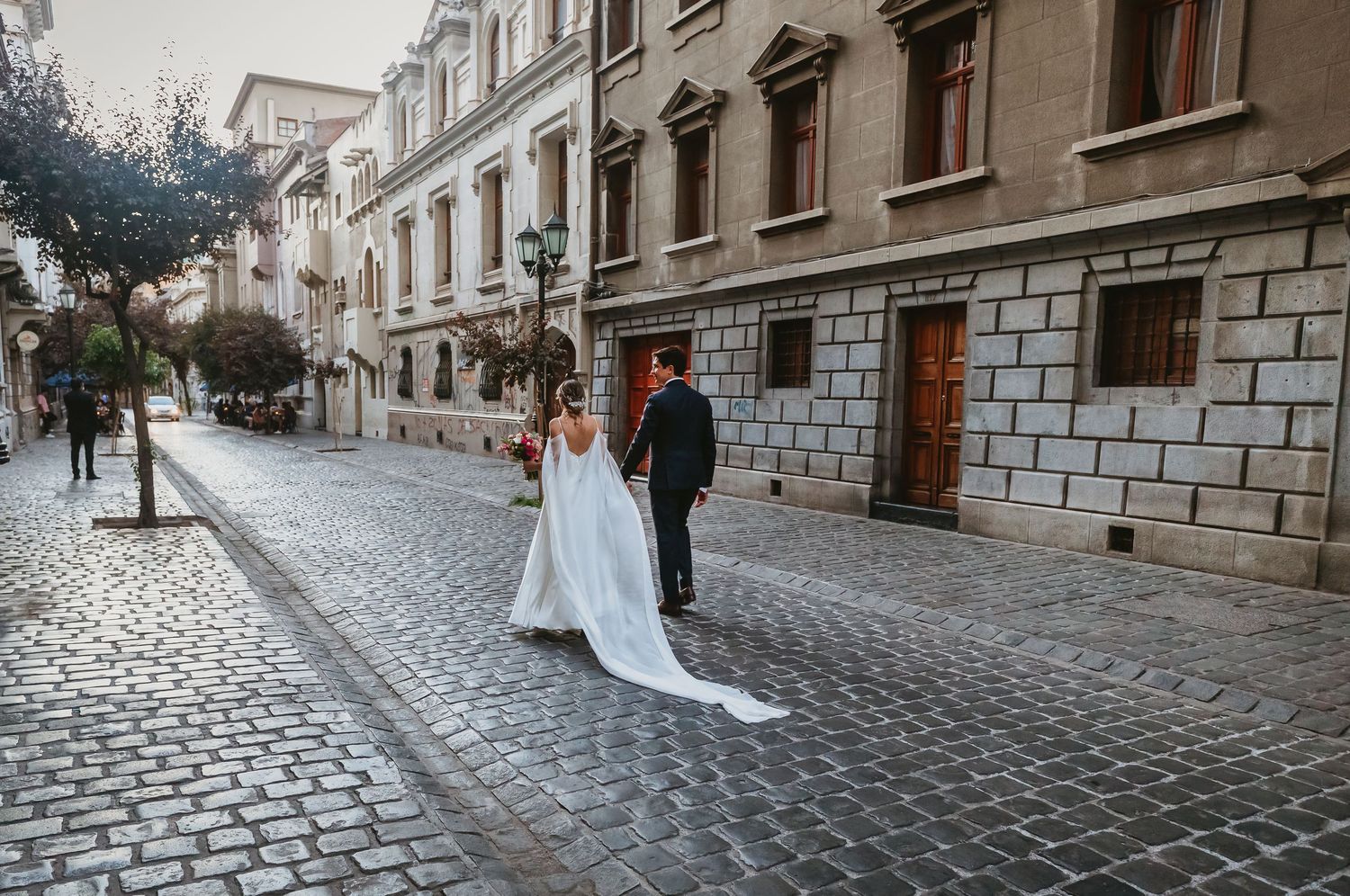 Bride and groom walking away on cobblestone street lined with historic buildings at dawn.