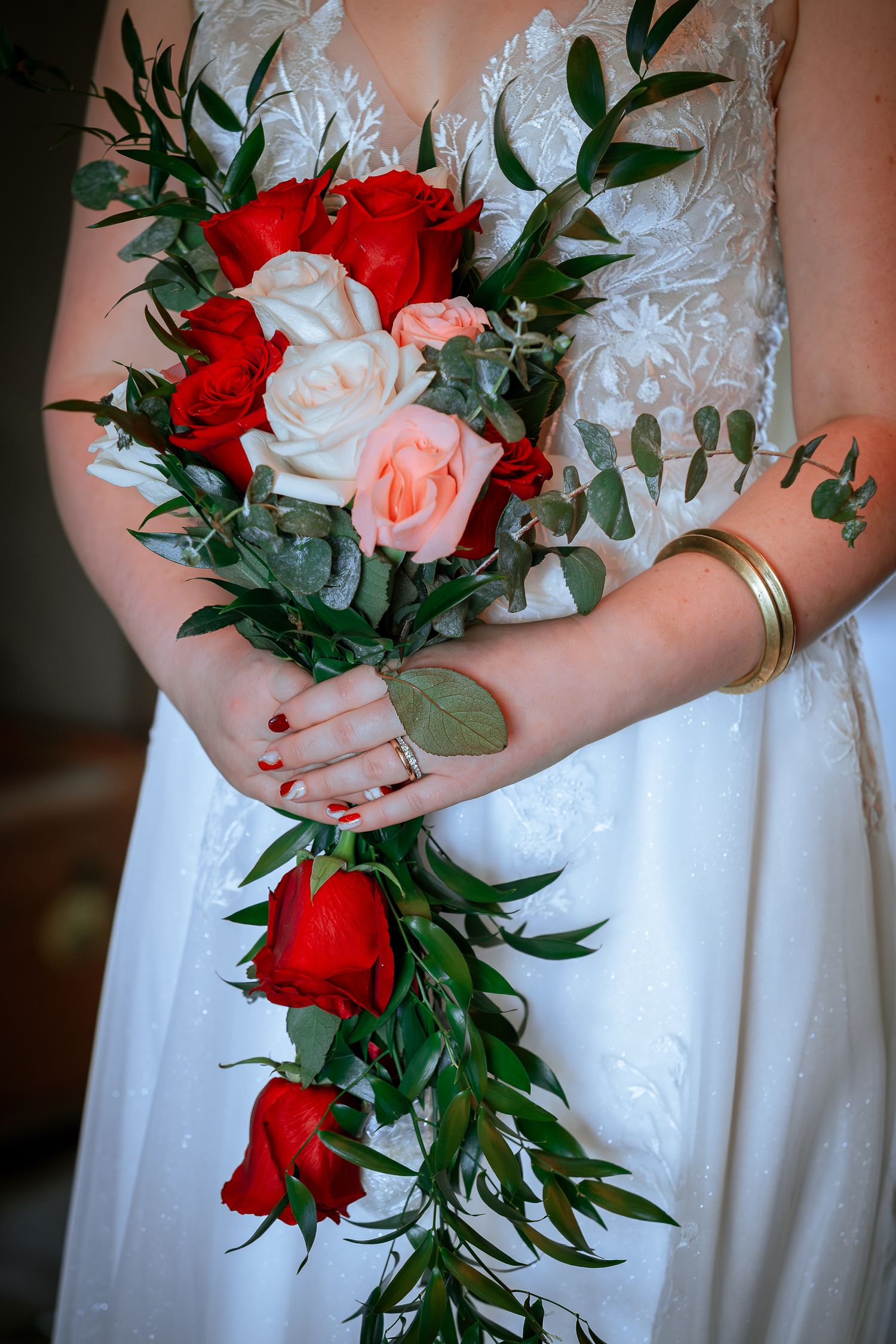 Bride in white lace gown holding a cascading bouquet of red, pink, and white roses with green foliage.