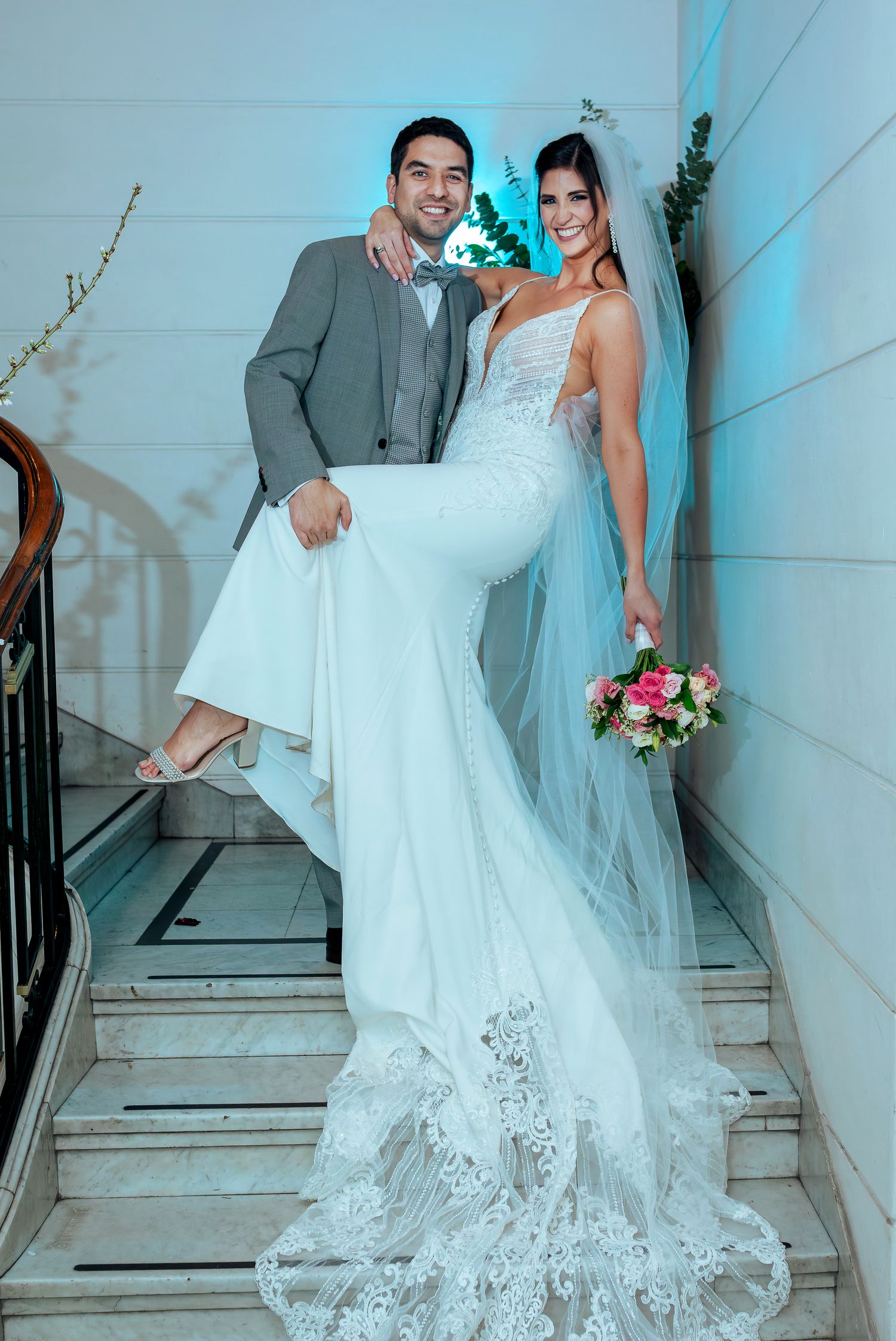 Bride in lace mermaid gown with veil holds bouquet beside groom in gray suit on marble staircase.