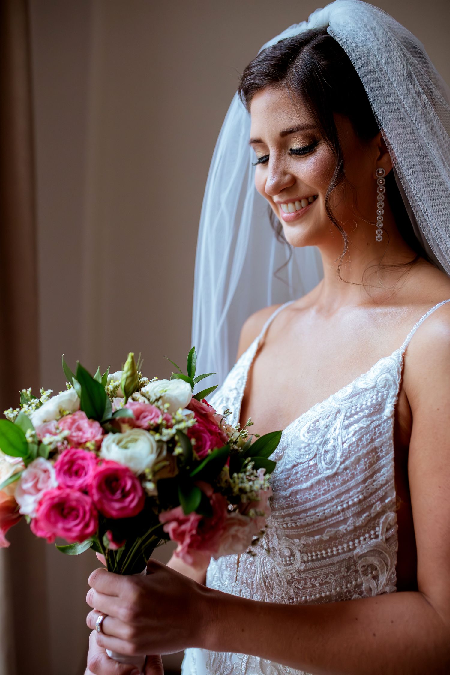 Smiling bride in white lace gown and veil holding a bouquet of pink and white roses on her wedding day.