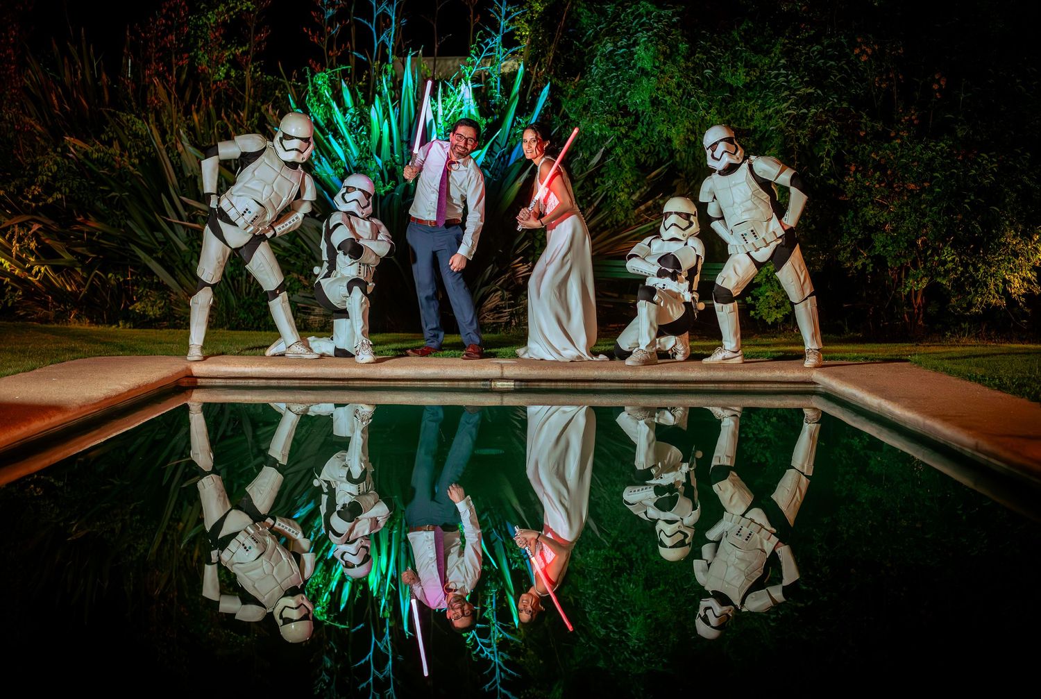 Bride and groom pose with Stormtroopers and lightsabers reflected in a pool at night.