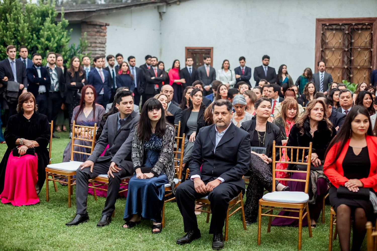 Wedding guests seated in gold chairs on garden lawn with large group portrait backdrop at outdoor ceremony.