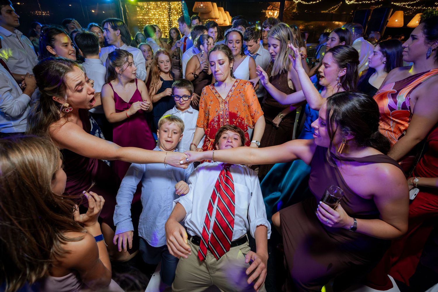 Energetic wedding reception dance floor with guests cheering around a man leaning back in a red tie.