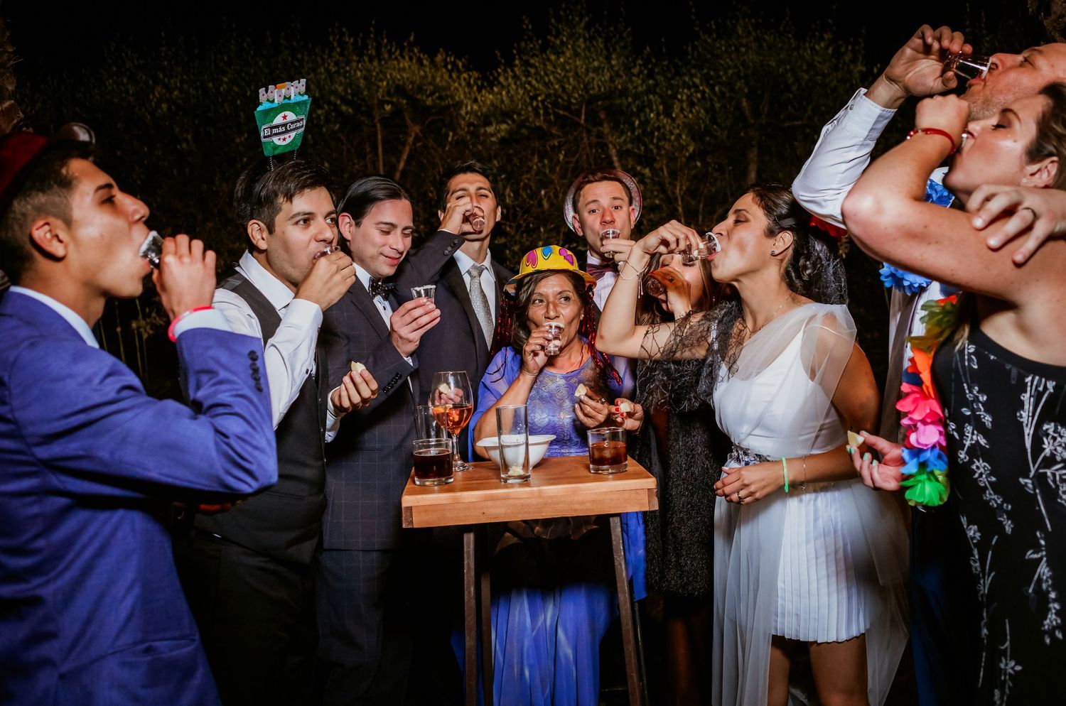 Wedding party guests take shots together around a small table during a lively outdoor wedding celebration at night.