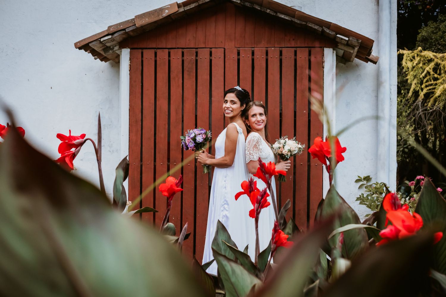 Two brides posing back-to-back with bouquets beside red barn door among lush red flowers.