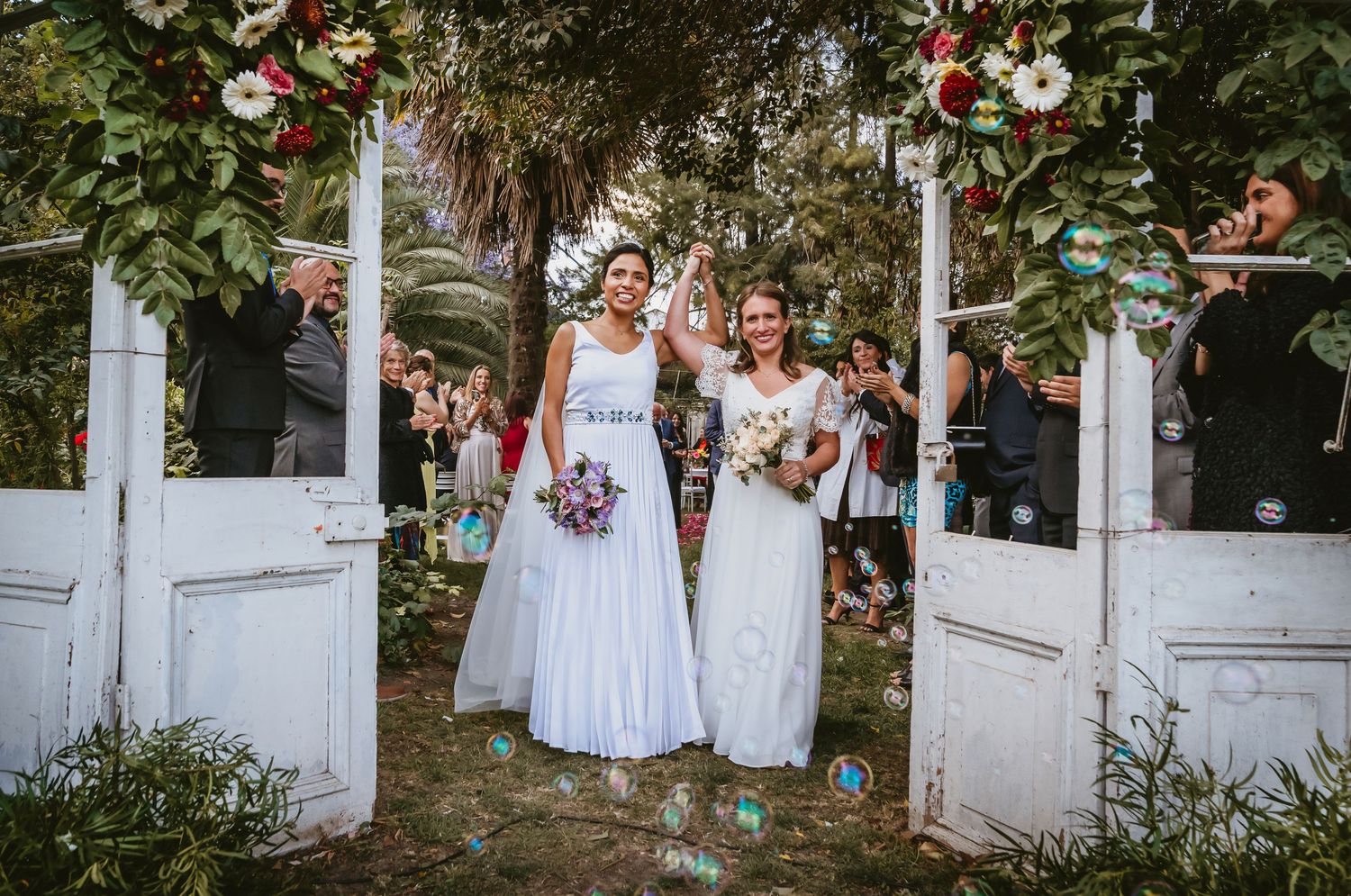 Two brides celebrate joyfully at a flower-adorned gate after their outdoor wedding ceremony.