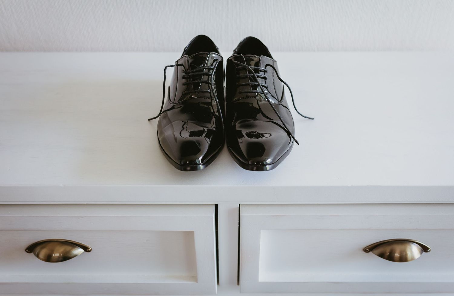 Pair of black patent leather Oxford dress shoes on white dresser drawer.
