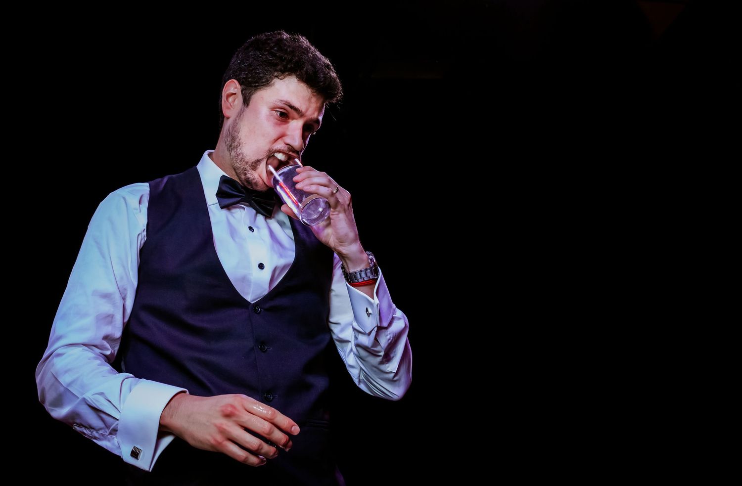 Elegant groom in navy vest sipping a drink against a dramatic dark background.